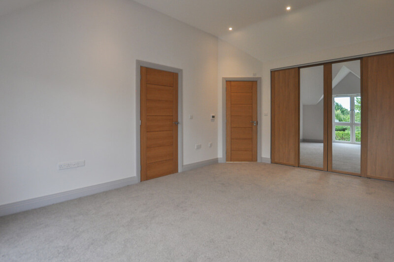 Spacious bedroom with white walls, warm grey carpet, large cupboard with mirrored centre doors.