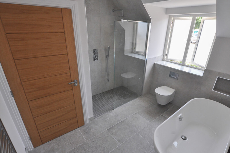 Premium bathroom with white suite including bath and shower, with large grey tiled walls and flooring and large wooden door.